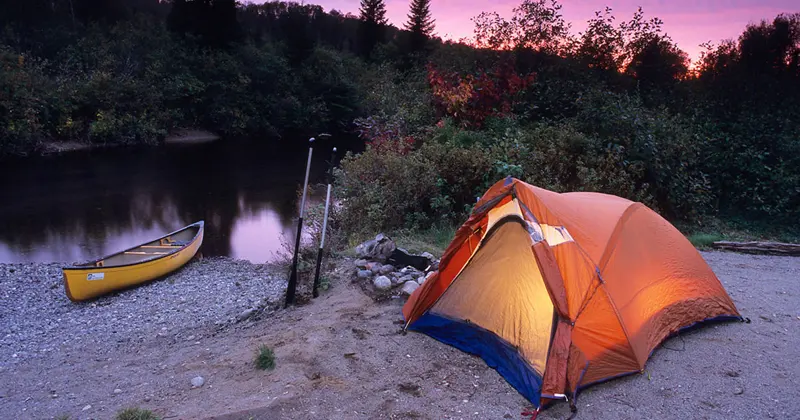 Orange tent and canoe on a river bank at dusk